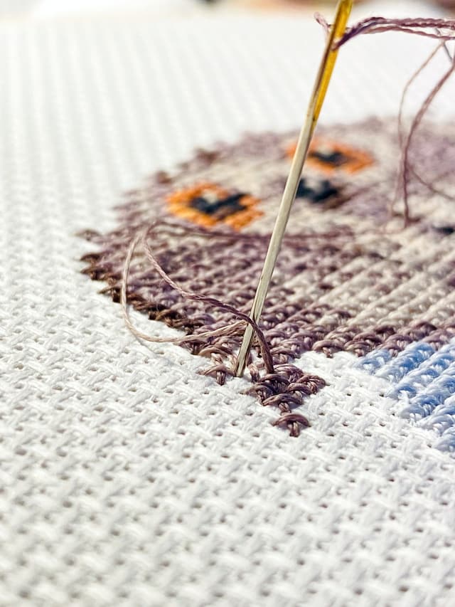 A needle pulling brown thread through a partly stitched cross-stitch design on white Aida fabric